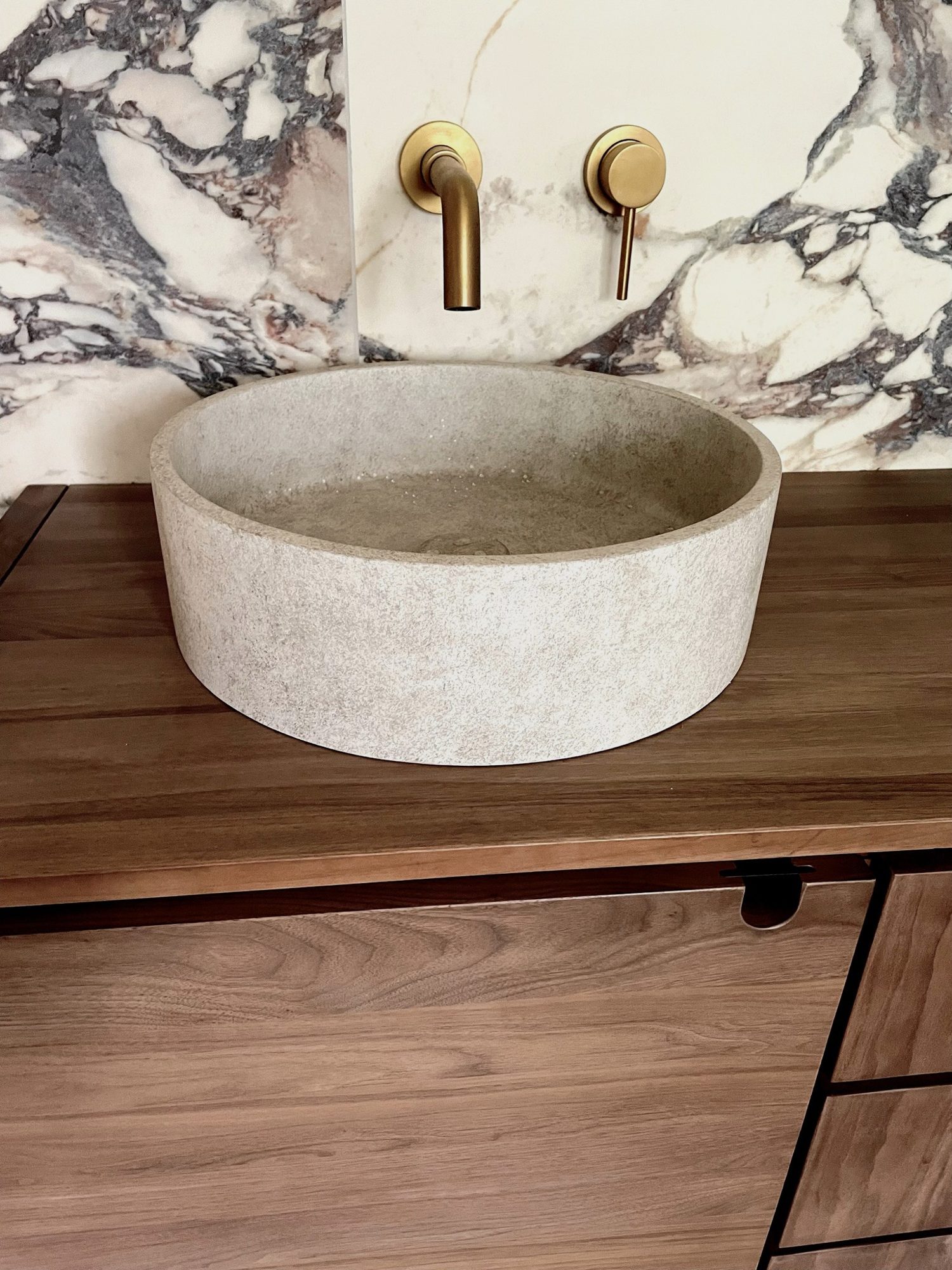 A round stone vessel basin on a walnut vanity, with brushed brass wall tap and controls against marble