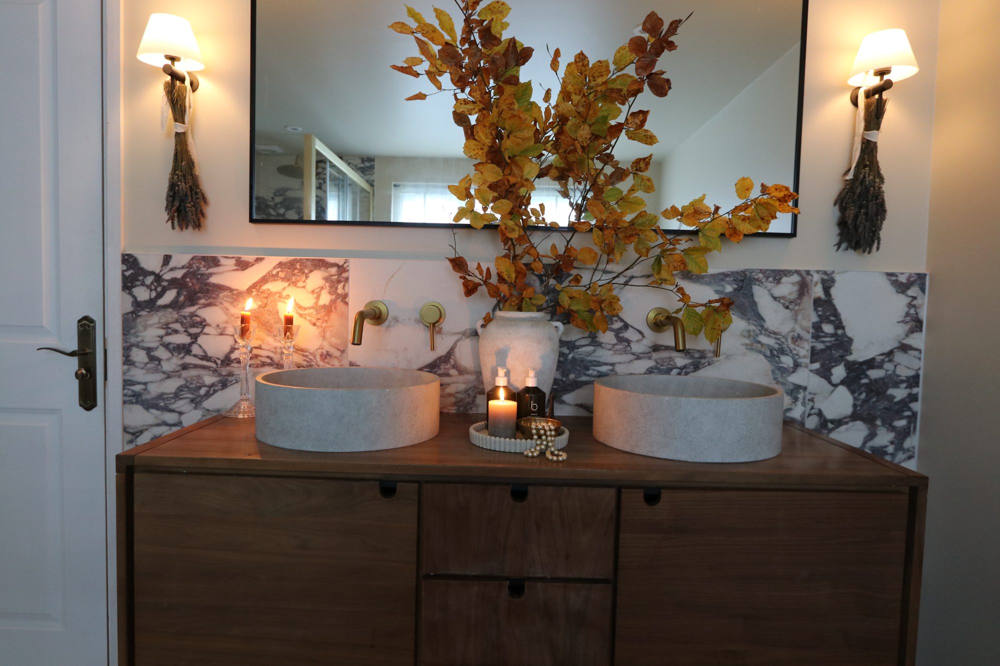 A marble bathroom with double stone basins on a walnut vanity, brass wall taps, sconces and autumn beech branches