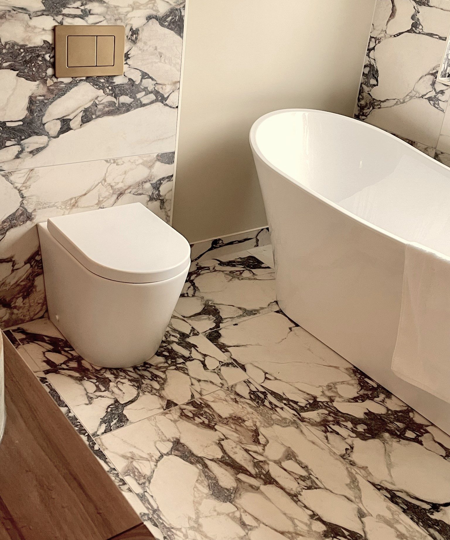 A freestanding white bath and wall-hung toilet in a fully marbled bathroom with a brass flush plate