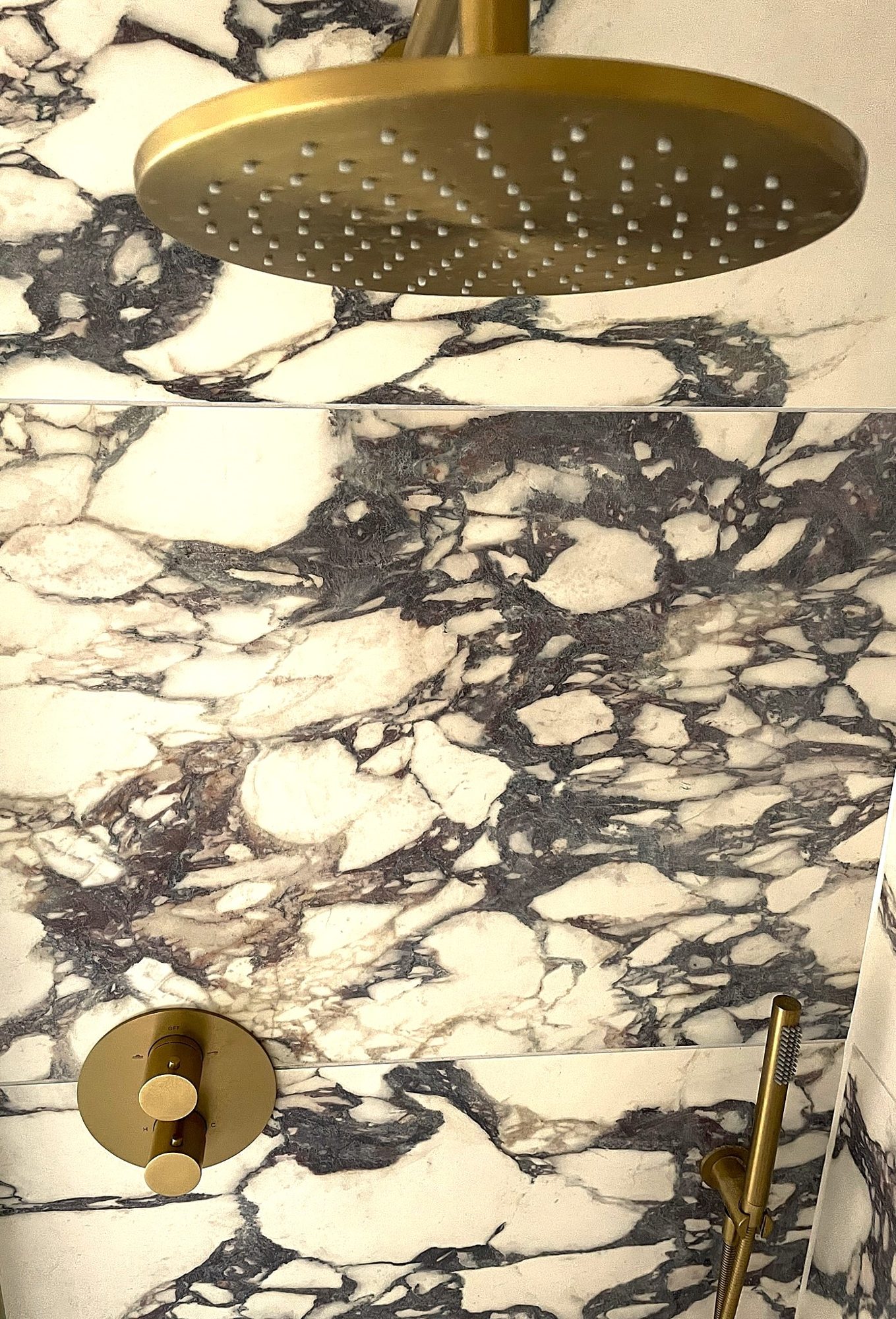 A brushed brass rainfall shower head and round thermostatic control against book-matched marble