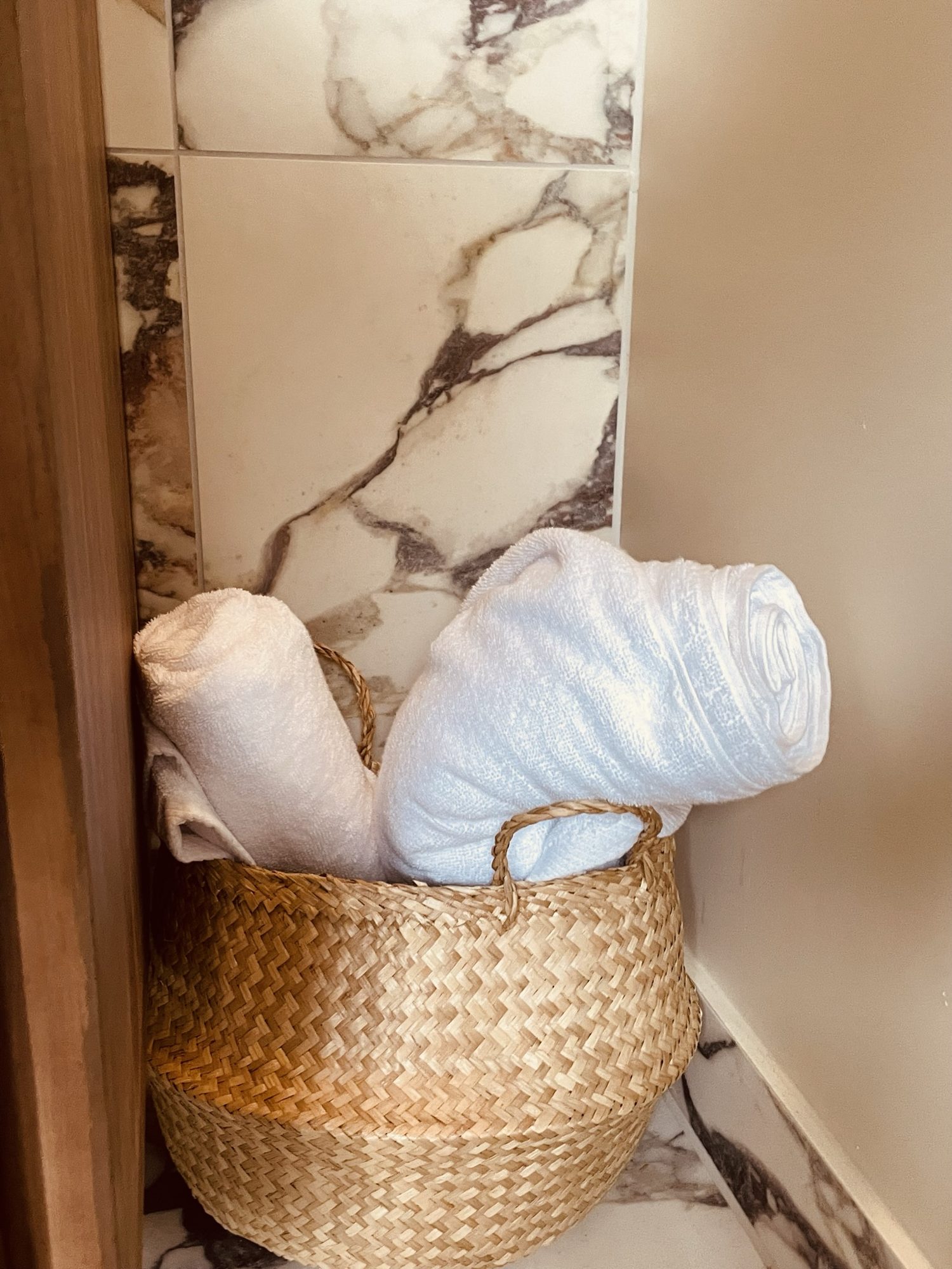 White towels rolled in a seagrass basket beside a walnut cabinet and marble wall