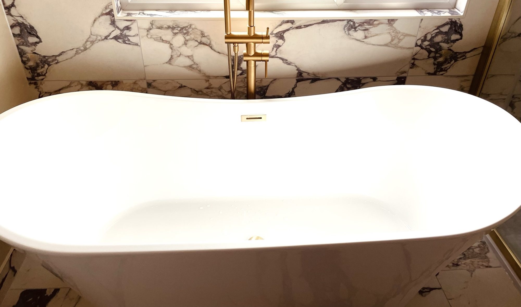 A white freestanding bath with a brushed brass floor-standing tap against marble