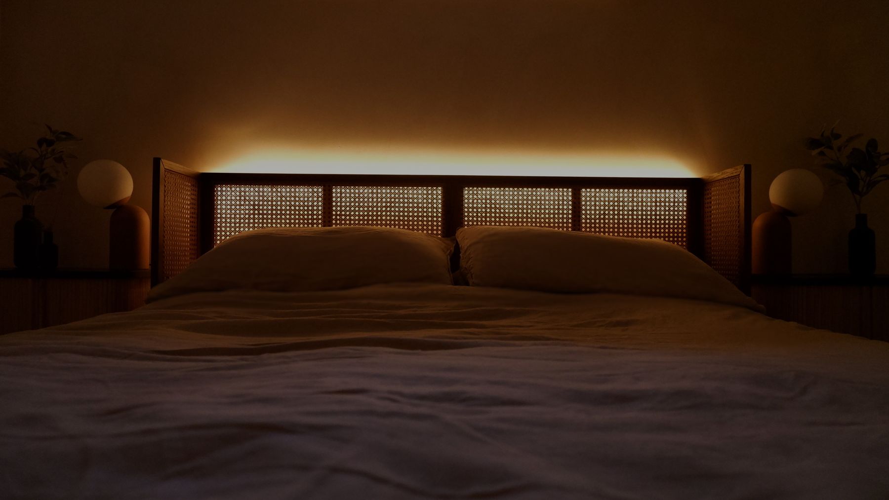 A bed with a cane headboard backlit by a warm strip of light, globe lamps either side