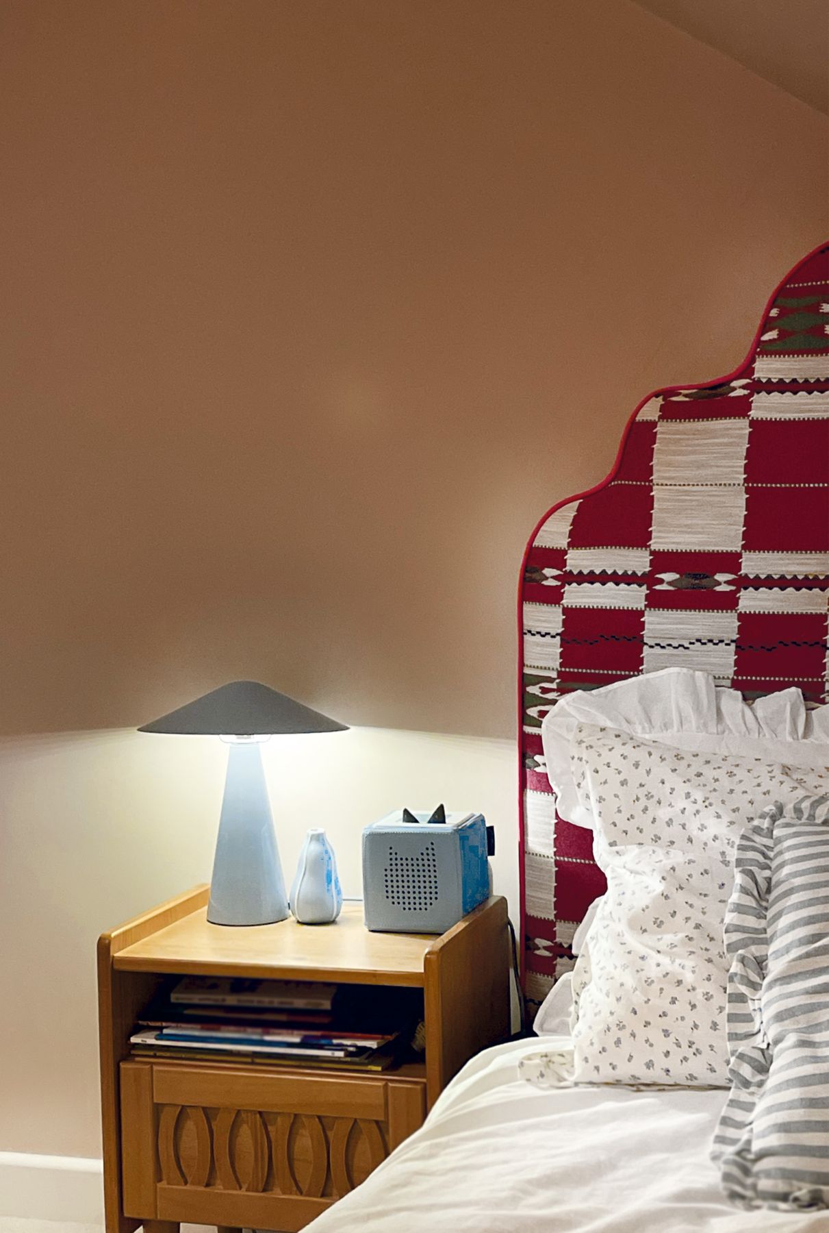 A child’s bed with a scalloped headboard in red and cream woven fabric, a blue mushroom lamp on a wooden bedside table