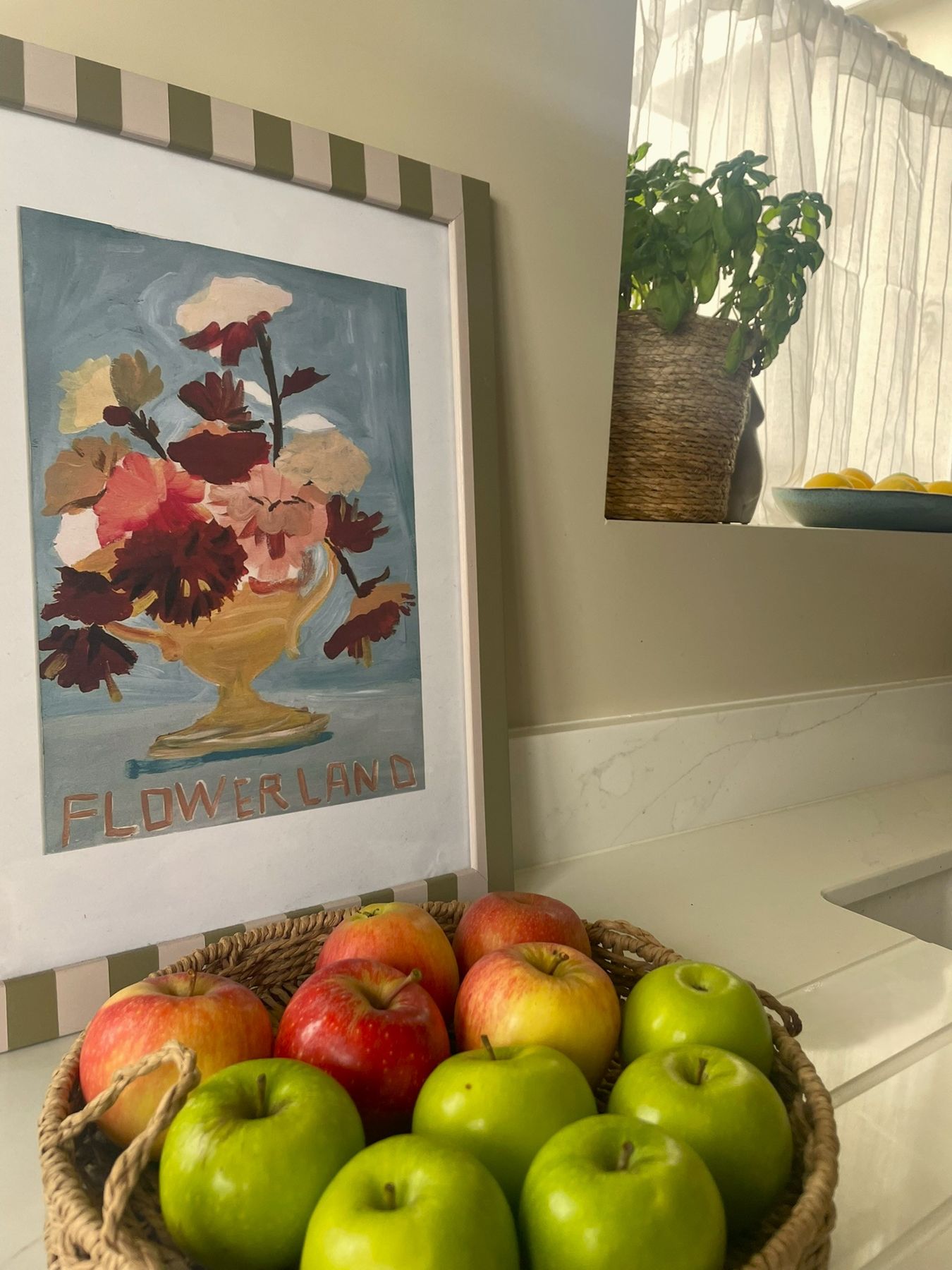 A bowl of green and red apples beneath a framed floral print