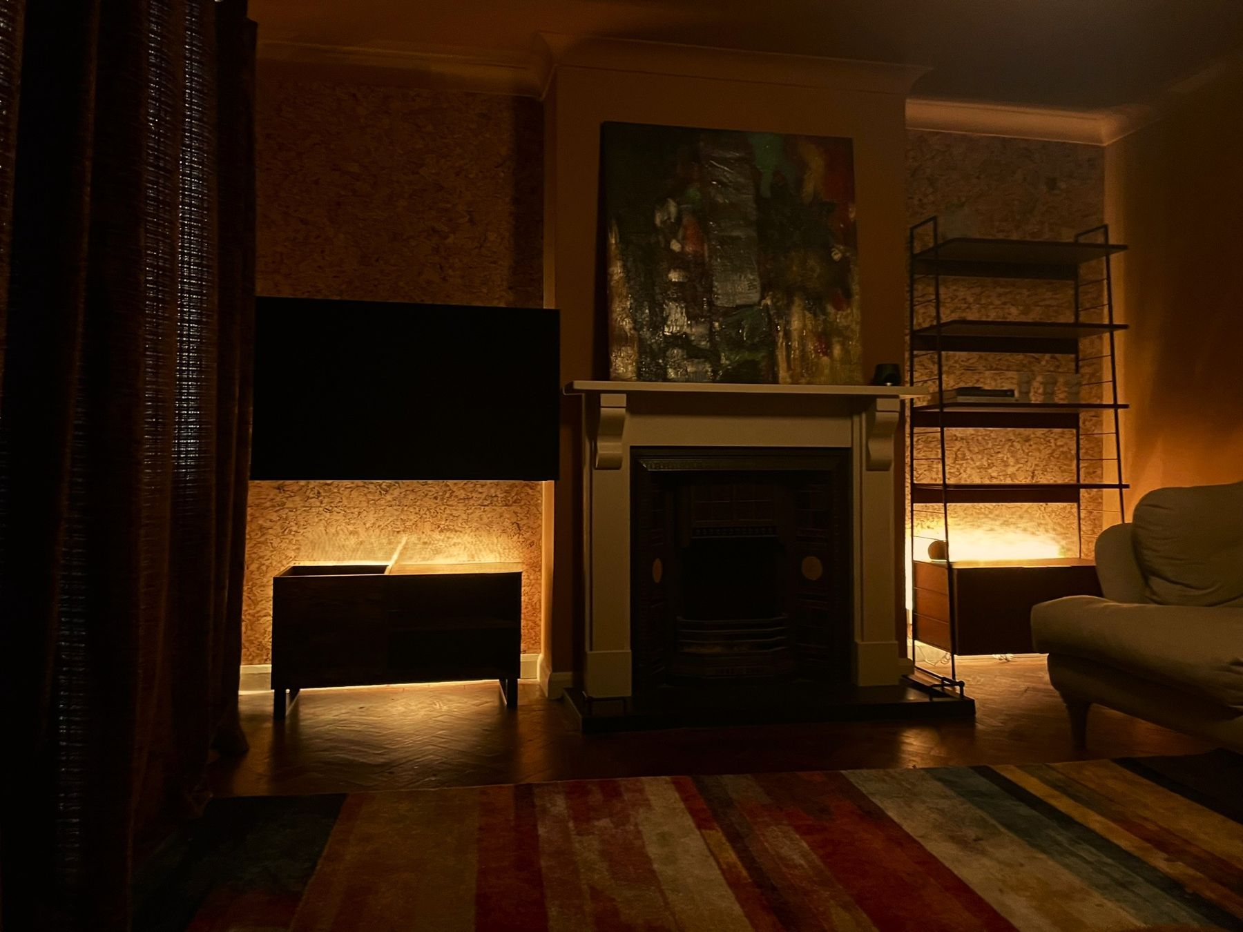 A sitting room at night: cork-lined alcoves lit from below, a cream fire surround, string shelving and a striped rug