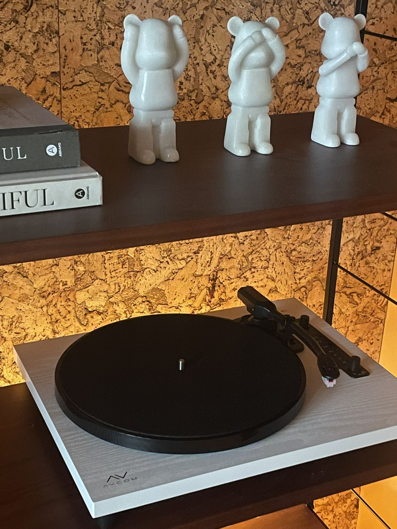 A white turntable below three white bear figures on a dark shelf, lit against cork