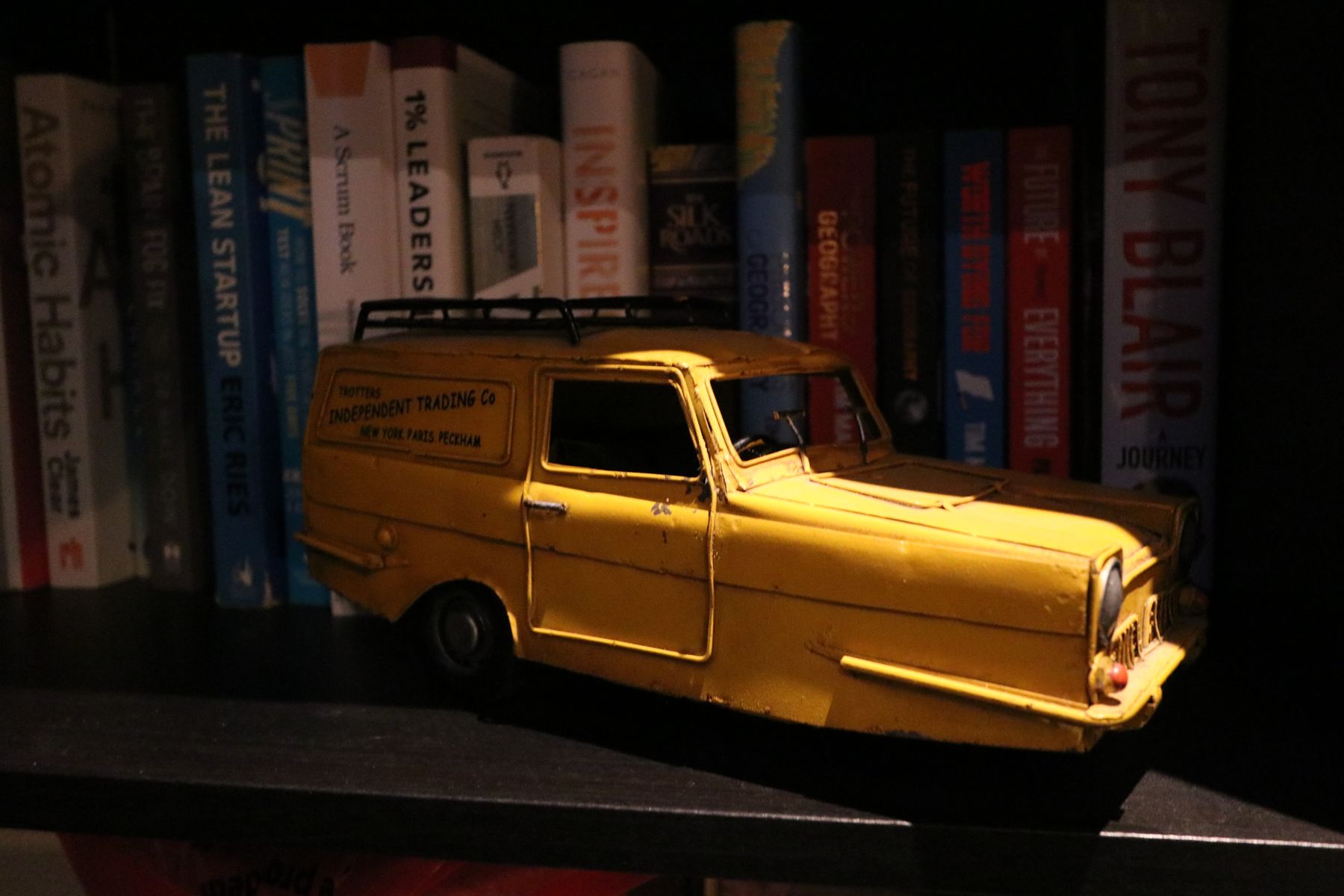A yellow model three-wheeler van in front of a row of books