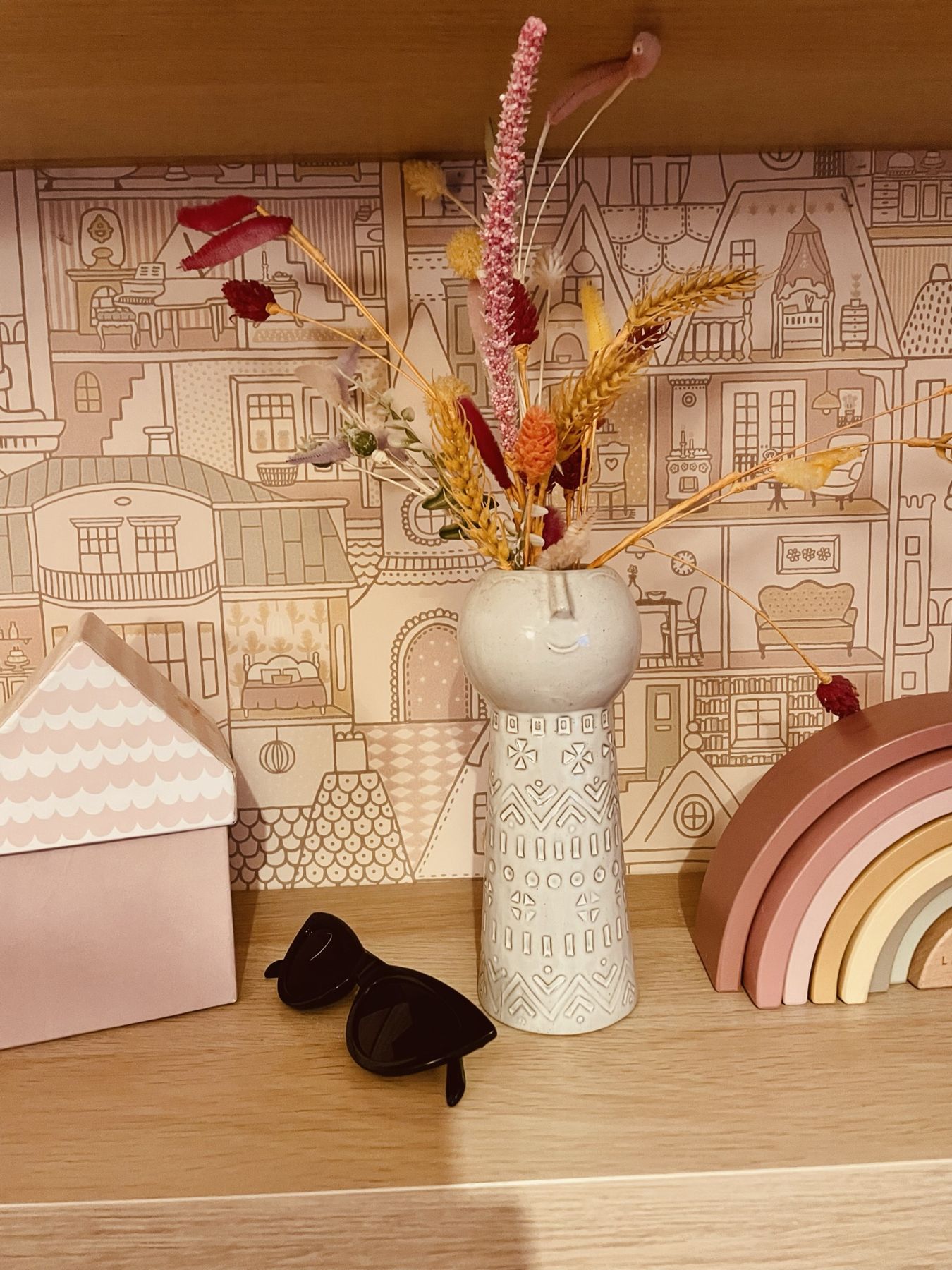 Wooden houses, a rainbow stacker and dried flowers in a white vase on a shelf