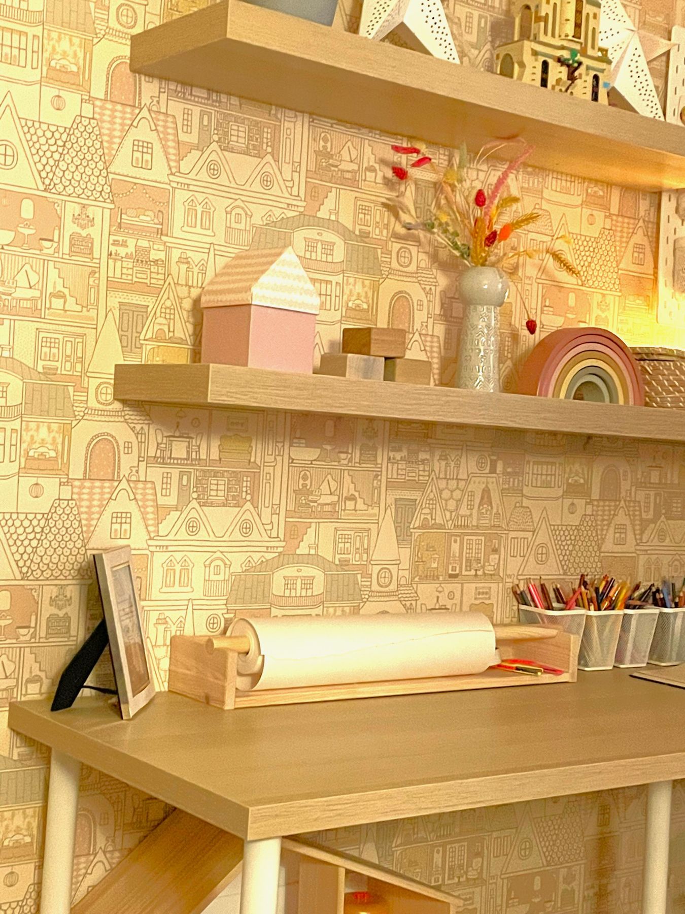 A pale wood desk and shelves against townhouse-print wallpaper, with wooden toys and dried flowers