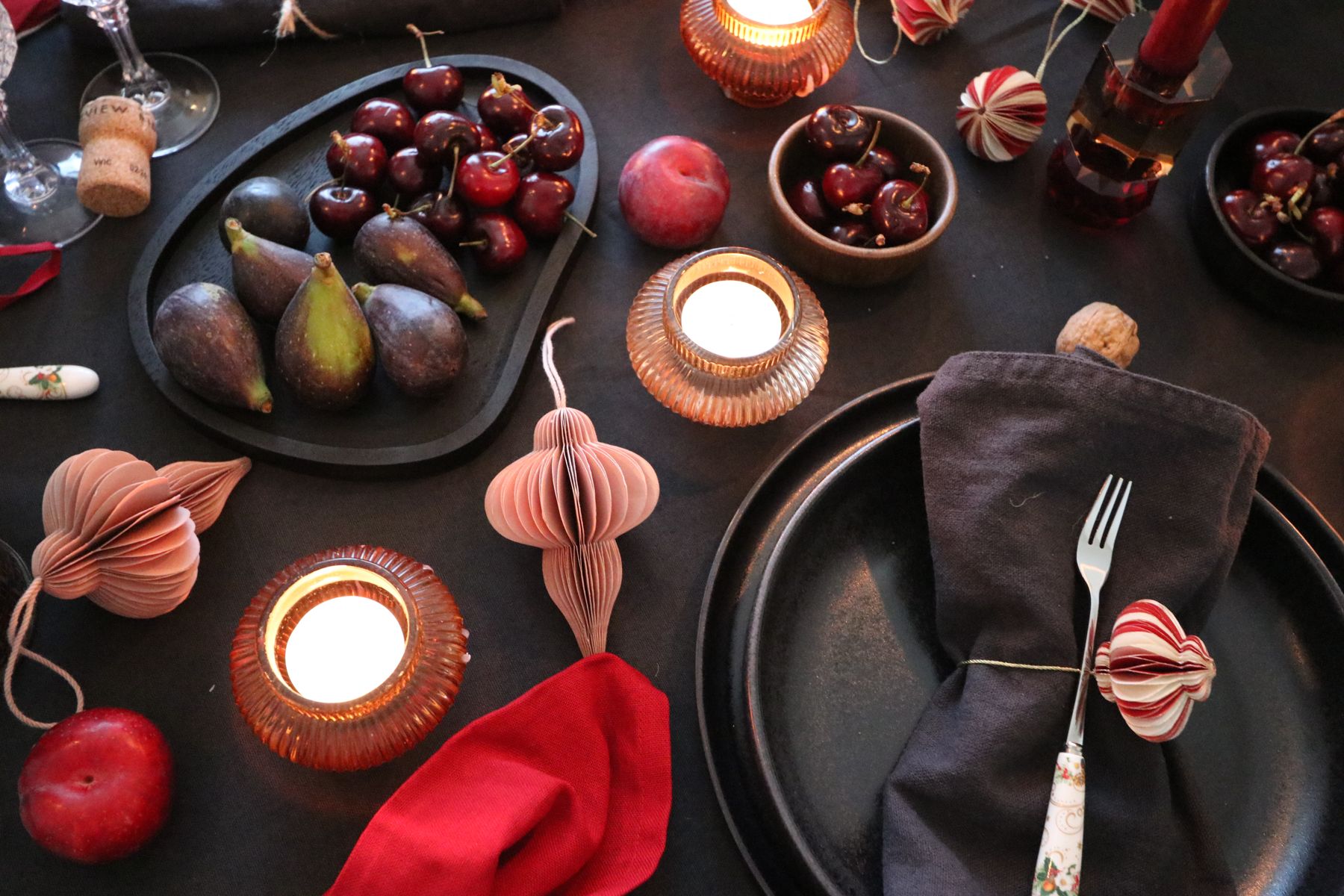 A dark Christmas table set with figs, cherries, pink paper baubles and glowing tealights
